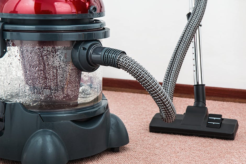 A yellow canister vacuum cleaner with a long black hose and metal wand attachment is positioned on a light-colored patterned area rug in a room with wooden flooring. The vacuum is turned off and not i